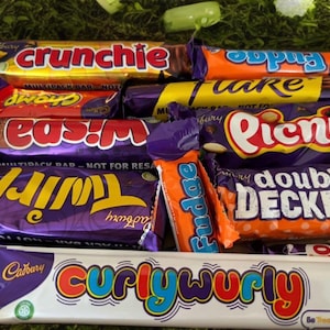 Pode incluir: Variedade de barras de chocolate Cadbury embaladas, incluindo Crunchie, Twirl e Curly Wurly. As embalagens coloridas apresentam várias fontes e designs. As barras estão dispostas sobre uma superfície verde.