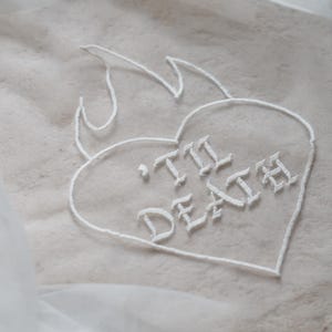 Könnte beinhalten: Nahaufnahme eines beigefarbenen Stoffes mit einem weißen, gestickten Herzdesign. Das Herz ist mit weißem Faden umrandet und trägt die Worte "'Til Death" in einer gotischen Schrift, mit Flammen über dem Herzen.