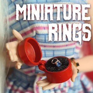 May include: A red ring box with a blue gemstone ring inside. The box is open and the ring is visible. The text "MINIATURE RINGS" is written in white letters on a pink and blue striped background.