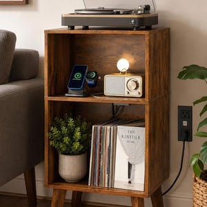 May include: A two-tier wooden nightstand with a dark brown wood grain finish and four angled legs. The top shelf holds a record player, a phone charging station, and a radio. The bottom shelf contains a potted plant and a book titled The Kinfolk Home.