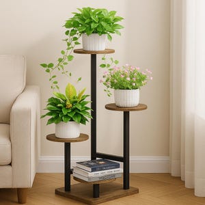 May include: A three-tiered plant stand with a dark brown wooden base and shelves, and black metal supports. The stand holds three white pots with green plants and pink flowers. Books with the titles "FASHION" and "WELCOME" are on the bottom shelf.