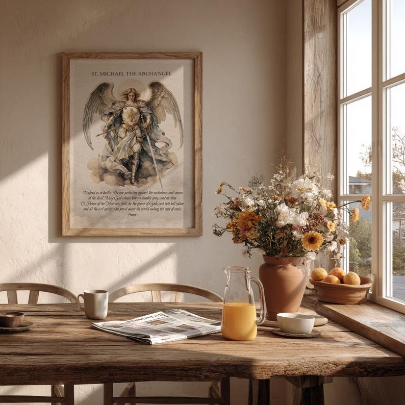May include: A framed print of St. Michael the Archangel hangs on a wall. The artwork depicts the archangel with wings, a shield, and a sword. The print is in a wooden frame. A wooden table is in the foreground with a newspaper, a mug, and a pitcher of orange juice.