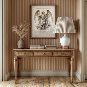 May include: A wooden console table with three drawers, a vase with dried plants, books, and a white lamp with a pleated shade. A framed print of St. Michael the Archangel hangs on the striped wall.