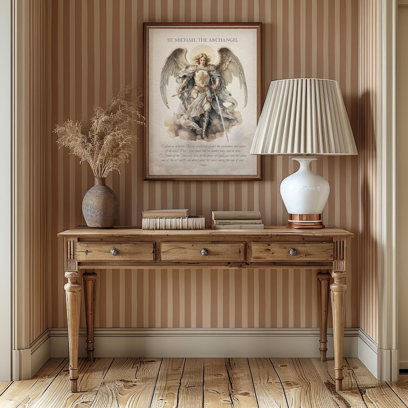 May include: A wooden console table with three drawers, a vase with dried plants, books, and a white lamp with a pleated shade. A framed print of St. Michael the Archangel hangs on the striped wall.