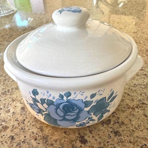May include: White ceramic casserole dish with a lid, featuring a blue rose floral design. The dish has two small handles and a knob on the lid. The floral pattern includes blue roses and green leaves.