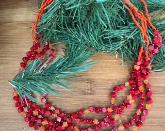 May include: A multi-strand necklace featuring red and orange beads. The necklace has a variety of bead shapes, including small round beads and larger square beads. The necklace is displayed on a wooden surface with green foliage.