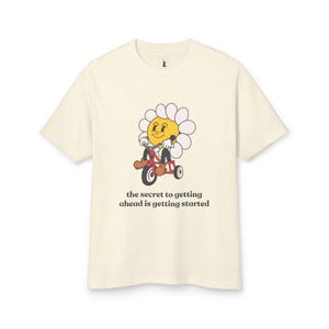 Könnte beinhalten: Ein cremefarbenes T-Shirt mit einer Cartoon-Gänseblümchen, die ein rotes Dreirad fährt. Die Gänseblümchen hat ein gelbes Gesicht und lächelt. Der Text "the secret to getting ahead is getting started" ist unter dem Bild gedruckt.