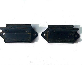 May include: Two black metal engine mounts with a rectangular shape and a raised lip on the top and bottom. The mounts have a hole on each side and a small lip on the front.