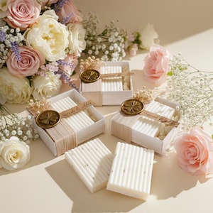 May include: White, rectangular soaps with vertical ridges are displayed in open white boxes, each tied with a beige ribbon and a gold seal. The boxes are arranged with a bouquet of pink and white roses and other flowers.