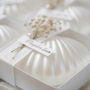 May include: A white, shell-shaped item in a white box, tied with a white ribbon. The box is decorated with small, dried flowers and a tag that reads "Regiden Lenn". The presentation is elegant and delicate, suitable for a gift.