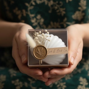 May include: A white seashell-shaped candle in a clear box, decorated with dried flowers and a gold wax seal. The seal has a leaf design and the text "Salt & Bush Co." The box is held in open hands.