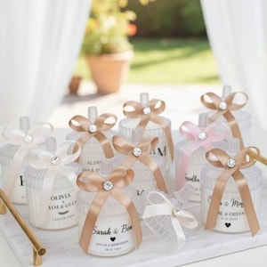 Puede incluir: Una colección de tarros de velas cilíndricos transparentes con tapas, adornados con cintas de raso y adornos de cristal. Los tarros están dispuestos sobre una bandeja de mármol blanco con asas doradas. Algunos tarros tienen etiquetas impresas con nombres y fechas. Las cintas son blancas, rosas y beige.