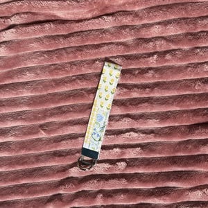 May include: A fabric keychain wristlet with a floral pattern. The wristlet features a yellow floral print, a blue and yellow checkered pattern, and a blue floral pattern. It has a silver metal ring for keys.