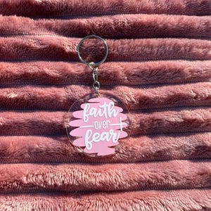 May include: A clear acrylic keychain with a silver ring and chain. The keychain features the words "faith over fear" in white script over a pink background. The background has a layered design. The keychain is set against a pink, textured background.
