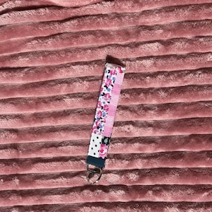 Puede incluir: Un llavero de tela con un llavero de metal. El llavero presenta un estampado floral, rayas rosas y un diseño de lunares. La pulsera mide aproximadamente 15 cm de largo.