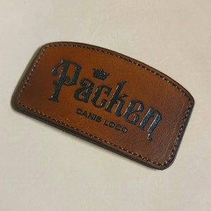 May include: A brown leather patch with black stitching and the text "Packen Canis Logo" in a stylized font with a crown above the word "Packen".