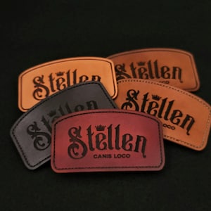 May include: Five leather patches with the word "Stellen" in a stylized font. The patches are in various colors, including brown, black, and red. One patch has the text "Canis Loco" below the word "Stellen".