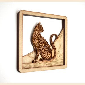 Puede incluir: Una pieza de arte de pared de madera con un dise&ntilde;o detallado e intrincado de un gato sentado en una roca. El gato est&aacute; tallado con patrones en espiral y la roca es texturizada. La pieza est&aacute; enmarcada en un simple marco de madera.
