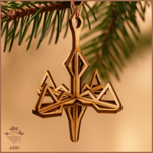 Op de afbeelding: Een houten geometrische kerstversiering met een gelaagd ontwerp, hangend aan een dun touwtje. De versiering heeft ruit- en driehoekvormen, met een natuurlijke houten afwerking. De achtergrond bevat een groene dennentak en een zachte bruine achtergrond.