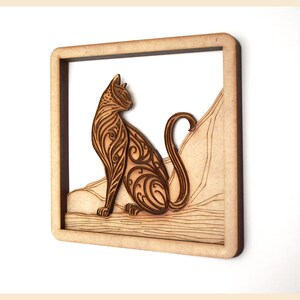 Puede incluir: Una pieza de arte de pared de madera con un gato estilizado con intrincados patrones. El gato est&aacute; sentado en una plataforma de madera con un fondo tallado.