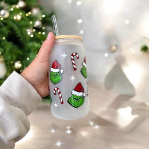 May include: A frosted glass reusable water bottle with a bamboo lid and a clear drinking straw. The bottle features a repeating pattern of green Grinch faces wearing Santa hats, candy canes, and white stars.