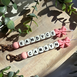 May include: Two personalised keychains with the names "KANDYCE" and "CAMRYN" spelled out in white letter beads. Each keychain features a pink bead, a pink bow, and a gold-coloured clasp. The keychains are displayed on a wooden tray with green foliage.
