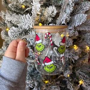 May include: Clear glass mug with a wooden lid, handle, and a clear straw. The mug is decorated with green Grinch faces wearing Santa hats and red and white candy canes. The background is a snowy Christmas tree.