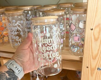 May include: A clear glass tumbler with a bamboo lid, featuring white printed text that reads Mama needs coffee, surrounded by decorative white bow and flower graphics. The glass is cylindrical with a smooth finish and a wide mouth opening.