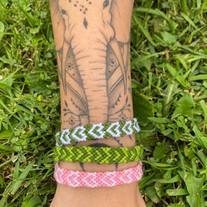 Może przedstawiać: Trzy bransoletki przyjaźni na nadgarstku osoby. Górna bransoletka jest biała i zielona z wzorem w chevron. Środkowa bransoletka jest zielona z wzorem w chevron. Dolna bransoletka jest różowa z wzorem w chevron.