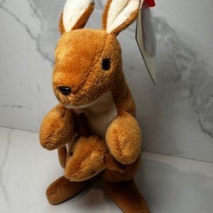 May include: A brown and white plush kangaroo toy with a joey in its pouch. The toy has a red and white tag attached to its ear.