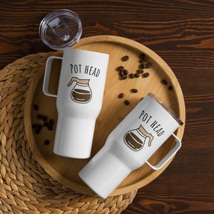 May include: Two white travel mugs with black text and a coffee pot graphic. The text on the mugs reads "Pot Head". The mugs are on a wooden tray with coffee beans scattered around them.
