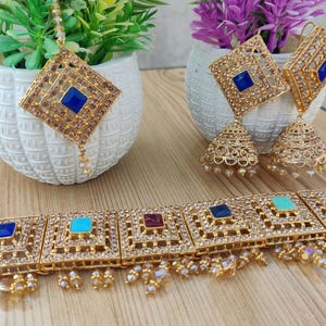 Puede incluir: Un conjunto de collar y pendientes de tono dorado con piedras azules, turquesa y rojas. El collar es de estilo gargantilla con cinco colgantes cuadrados. Los pendientes son de estilo colgante con un colgante cuadrado y un dije en forma de campana.