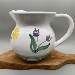May include: White ceramic pitcher with a rounded body and a curved handle. The pitcher is decorated with hand-painted yellow and purple floral designs with green leaves. The pitcher sits on a wooden cutting board.