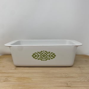May include: A rectangular, white baking dish with a green decorative design in the center. The dish is sitting on a wooden surface. The dish has a simple, classic design.