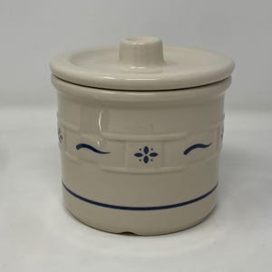 May include: Cream-colored ceramic jar with a lid. The jar features a blue band at the base and a decorative pattern of blue curved lines and floral designs. The lid has a small knob for easy removal.