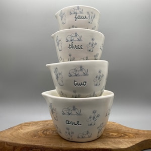 Easter scene ceramic measuring cups by Rae Dunn. Measurements in cursive on front. Sky blue and white in color. Adorable for use or display