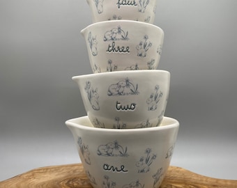 Easter scene ceramic measuring cups by Rae Dunn. Measurements in cursive on front. Sky blue and white in color. Adorable for use or display