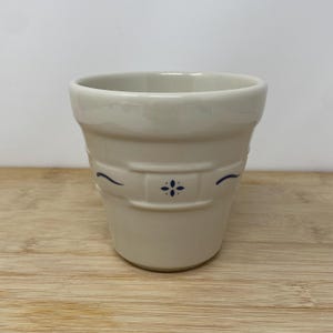 May include: A cream-colored ceramic planter with a flared rim and a tapered body. The exterior features a raised rectangular panel detail centered with a small, dark blue floral motif and flanked by two thin, curved dark blue decorative lines.