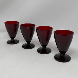 May include: Four burgundy-colored stemmed glasses with a conical bowl and a rounded base. The glasses have a deep red interior and a dark exterior, creating a striking contrast. The glasses are arranged on a white surface.