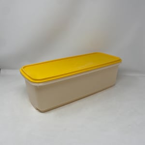 May include: A rectangular food storage container with a yellow lid and a beige base. The lid is flat and fits snugly on the container. The container is made of durable plastic and is designed for storing food items.