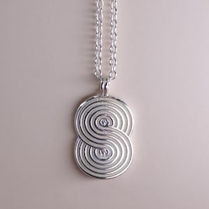 Pode incluir: Um colar de prata com um pingente em forma de símbolo de infinito. O pingente é feito de múltiplos círculos concêntricos.