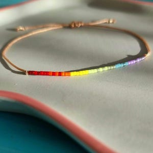 Peut inclure: Un bracelet délicat composé d'un cordon fin beige et d'une rangée de petites perles cylindriques disposées en dégradé arc-en-ciel, allant du rouge et orange au jaune, vert, bleu et violet, doté d'un fermoir à nœud coulissant réglable.