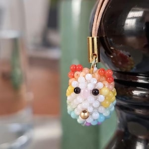 Könnte beinhalten: Ein farbenfroher Perlenanhänger in Form eines Tierkopfes. Der Anhänger hat ein Regenbogenmuster mit Farben wie Rot, Orange, Gelb, Weiß, Blau und Lila. Er hat schwarze Perlenaugen und eine goldene Perlnase. Der Anhänger ist an einer beigefarbenen Kordel befestigt.