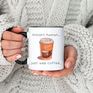 Może przedstawiać: Biały kubek do kawy z czarnym rantem i uchwytem. Kubek ma kresk&oacute;wkową ilustrację kubka do kawy z tekstem "Instant human... Just add coffee..."