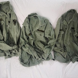 May include: Three olive green long-sleeve military-style jackets with collars, button-front closures, and two chest pockets. Each jacket features a black patch on the left shoulder and one jacket displays the text U.S. ARMY above the pocket.