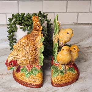 May include: A pair of ceramic bookends depicting a hen and chicks. The hen is brown and tan with red details, and the chicks are yellow. The bookends have a basket weave pattern and are adorned with leaves and berries.