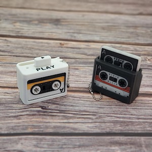 May include: Two retro-style cassette tape keychains. One is white with "PLAY" printed on it, and the other is black. Both have a silver chain. The cassette tapes have a retro design with orange, red, and white details.