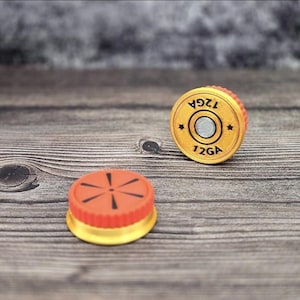 May include: Two bottle stoppers with a shotgun shell design. One stopper is orange and gold with a black design. The other is gold with the text "12GA" and a star design. The stoppers are on a wooden surface.