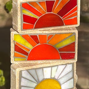 Puede incluir: Tres piezas de arte de mosaico rectangulares apiladas. Cada pieza presenta un diseño de sol con rayos radiantes. Las dos superiores tienen tonos naranja, rojo y amarillo, mientras que la pieza inferior tiene un diseño de sol blanco y amarillo.