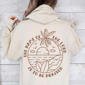 Puede incluir: Sudadera con capucha beige con un gráfico marrón en la espalda. El gráfico presenta una palmera, sol, olas y el texto "THE NAME OF THE LORD IS TO BE PRAISED" en un diseño circular. La sudadera tiene capucha y mangas largas.